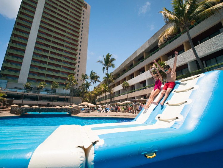 Boy and girl sliding down inflatable water slide with palm trees and resort in background