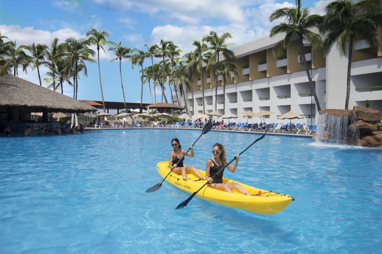 Two women in yellow kayak paddling in pool with lounge area and palm trees in background