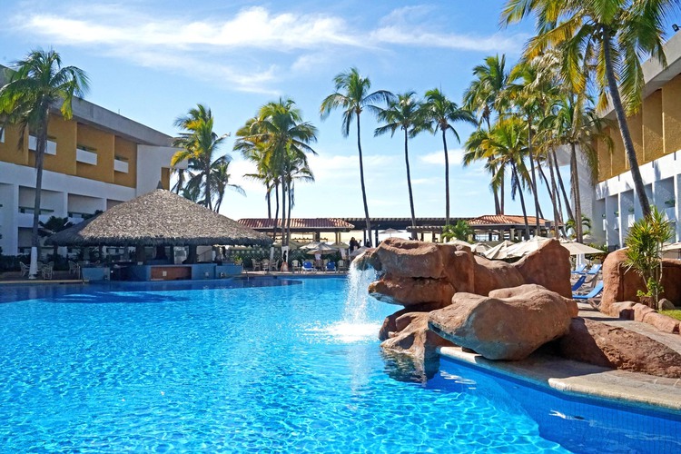 Pool area with waterfall over rocks and palapa bar with palm trees in background